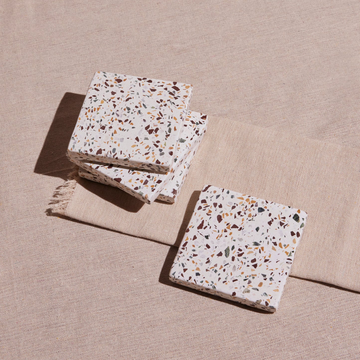 A set of four terrazzo coasters with a multicolored pattern, placed on a beige fabric surface.