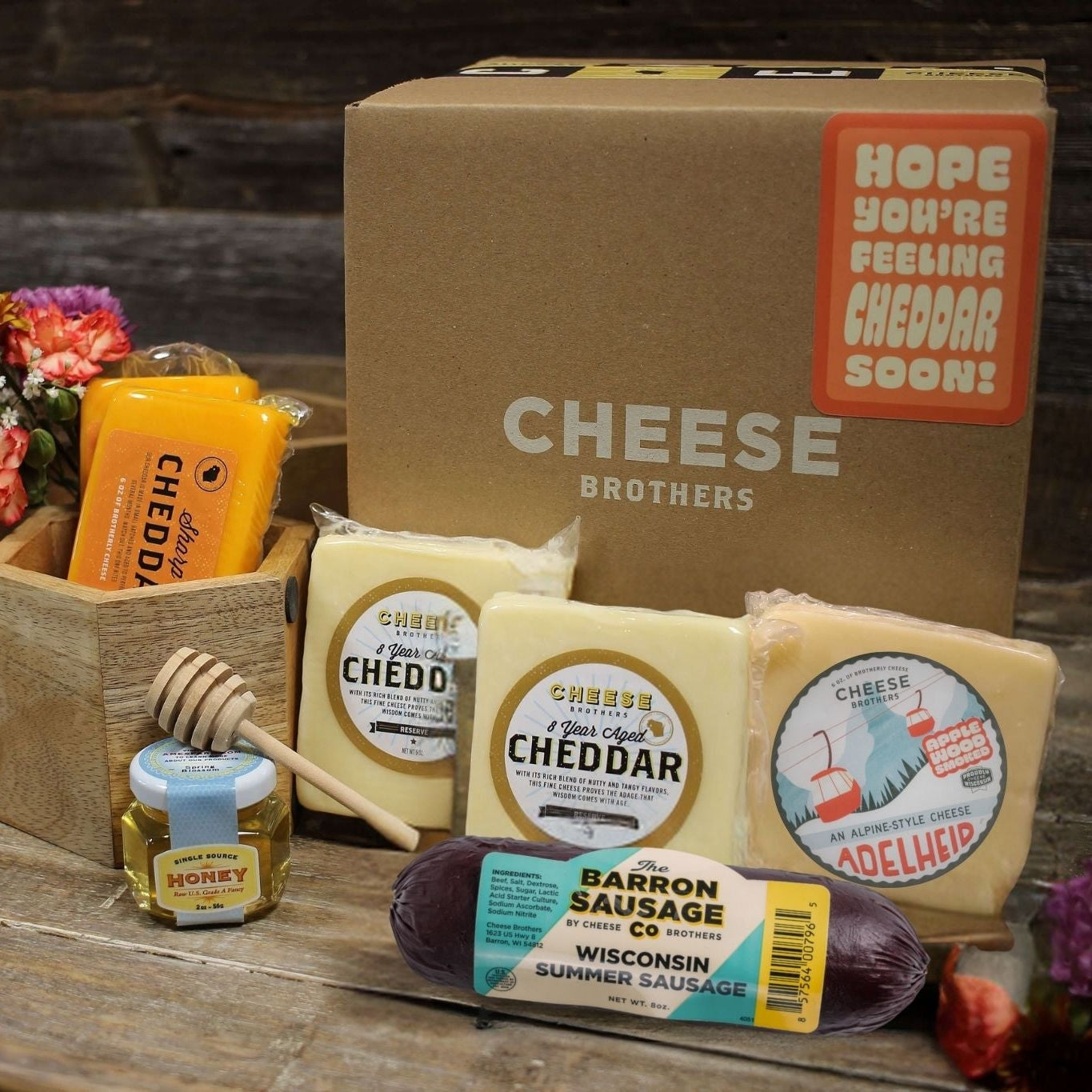 Cheese and sausage gift set with flowers and a box labeled 'Hope You're Feeling Cheddar Soon!' on a wooden surface.