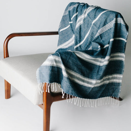A navy and natural striped throw blanket draped over a modern wooden armchair with a cushion on it, set against a neutral background.