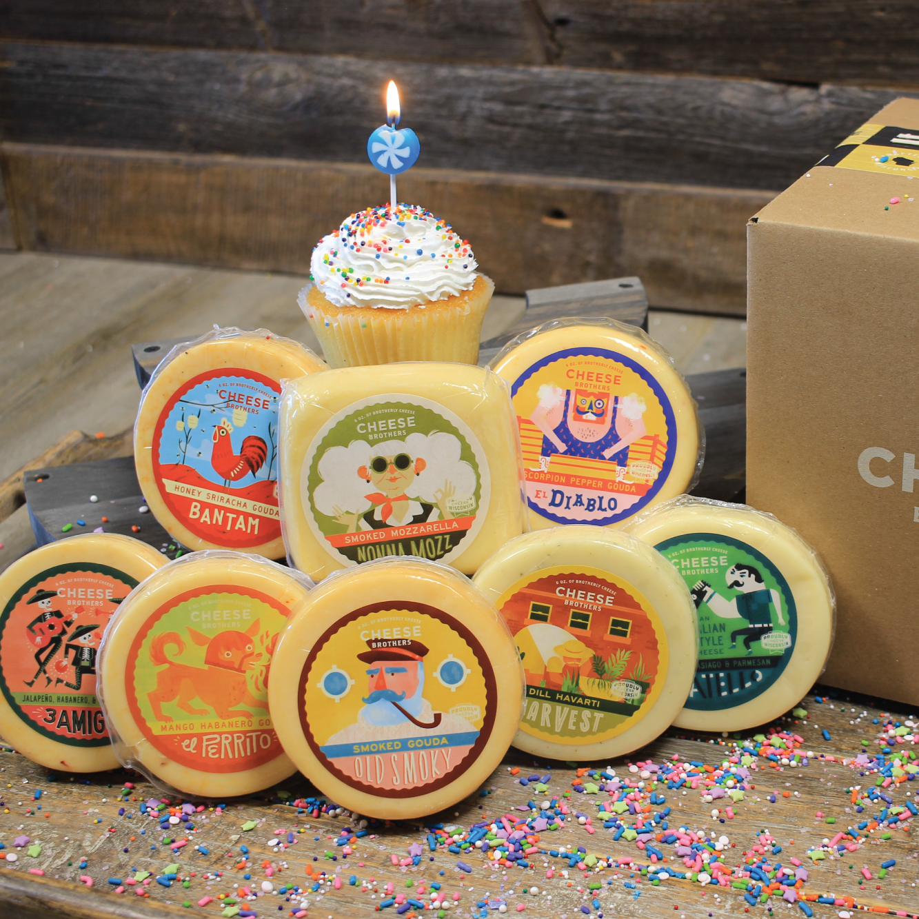 Collection of cheese wheels with colorful labels and a birthday cake with candle next to a cardboard box labeled 'Cheese Brothers' on a rustic wooden surface.