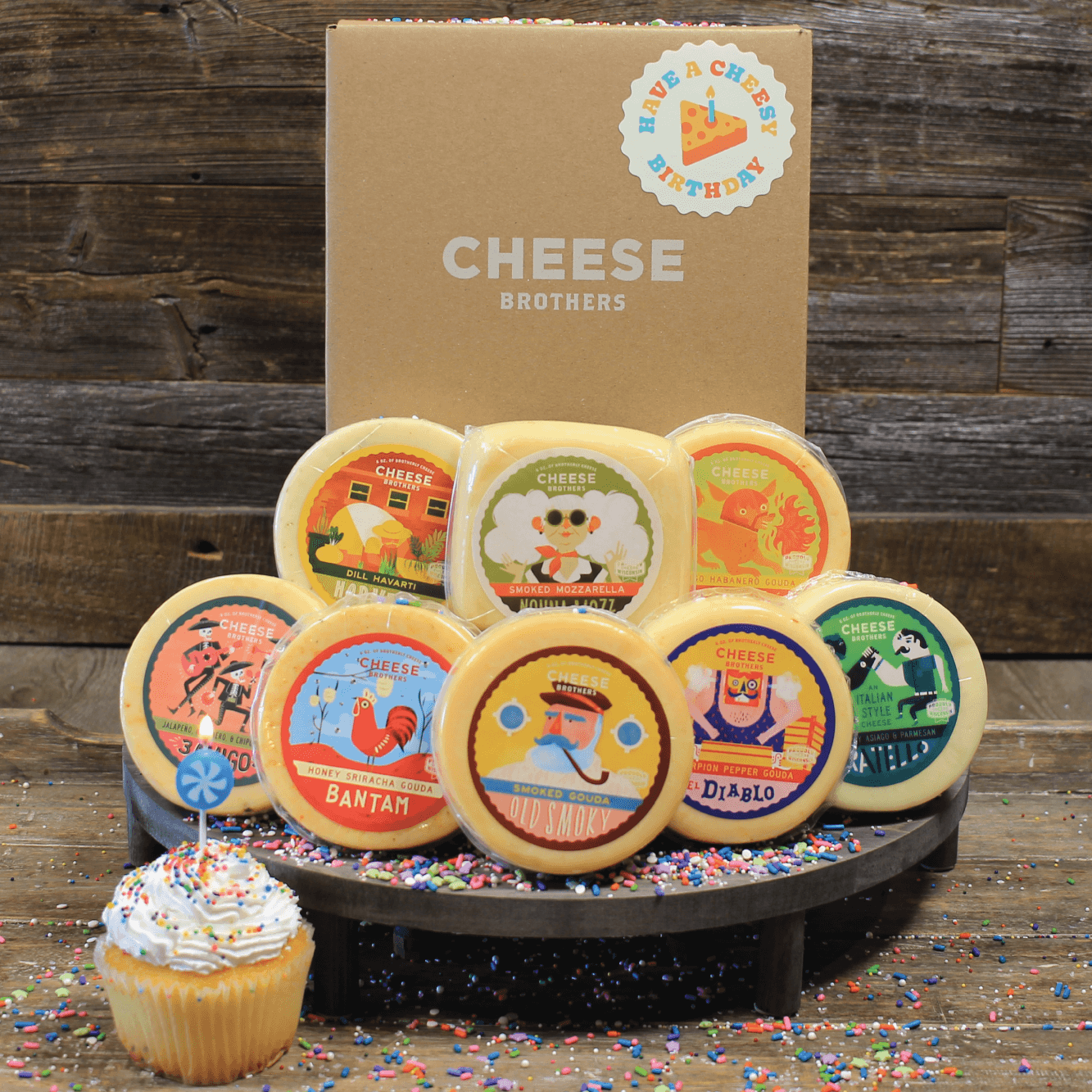 Assorted cheese wheels with colorful labels on a wooden stand, against a wooden background with 'Cheese Brothers' branding.